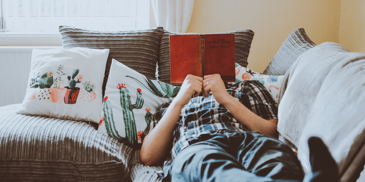 A man relaxing on a couch and holding a book in front of his face, reading.