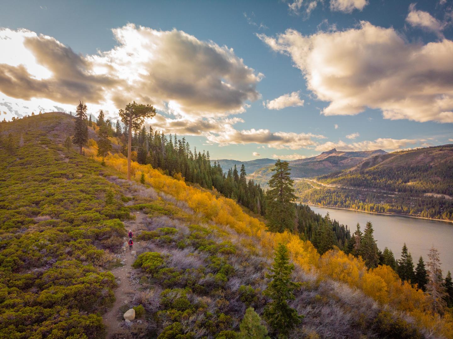 Hiker on a trail through colorful autumn trees by a lake under a partly cloudy sky.