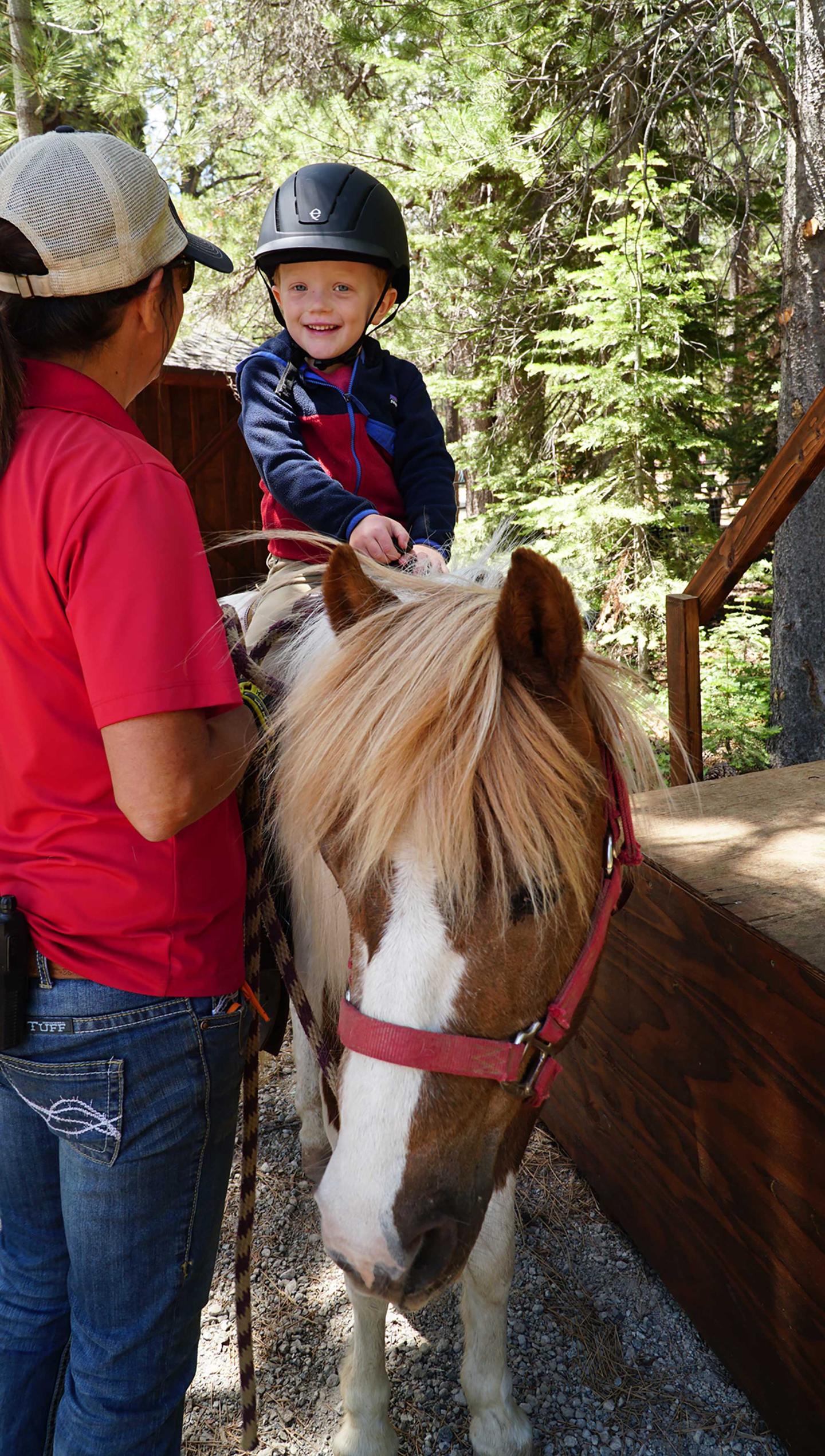 Child smiling on a pony with a guide in a forest setting.