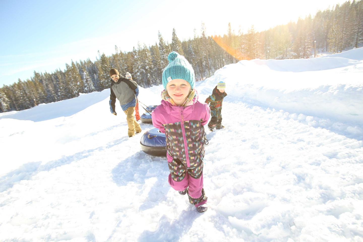 Child in snowsuit walking on snowy hill, bright sunlight, trees in background.