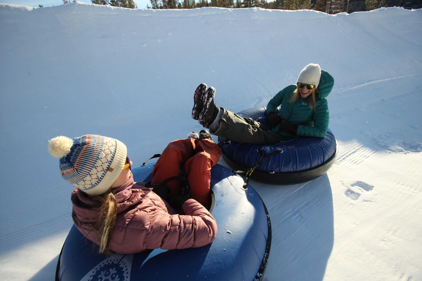 Two people tubing on snow in winter clothing, sunny day.