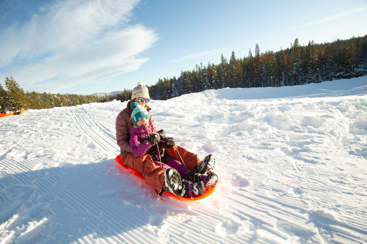 Sleigh ride on snow, adult and child bundled up, sunny day, forest backdrop.