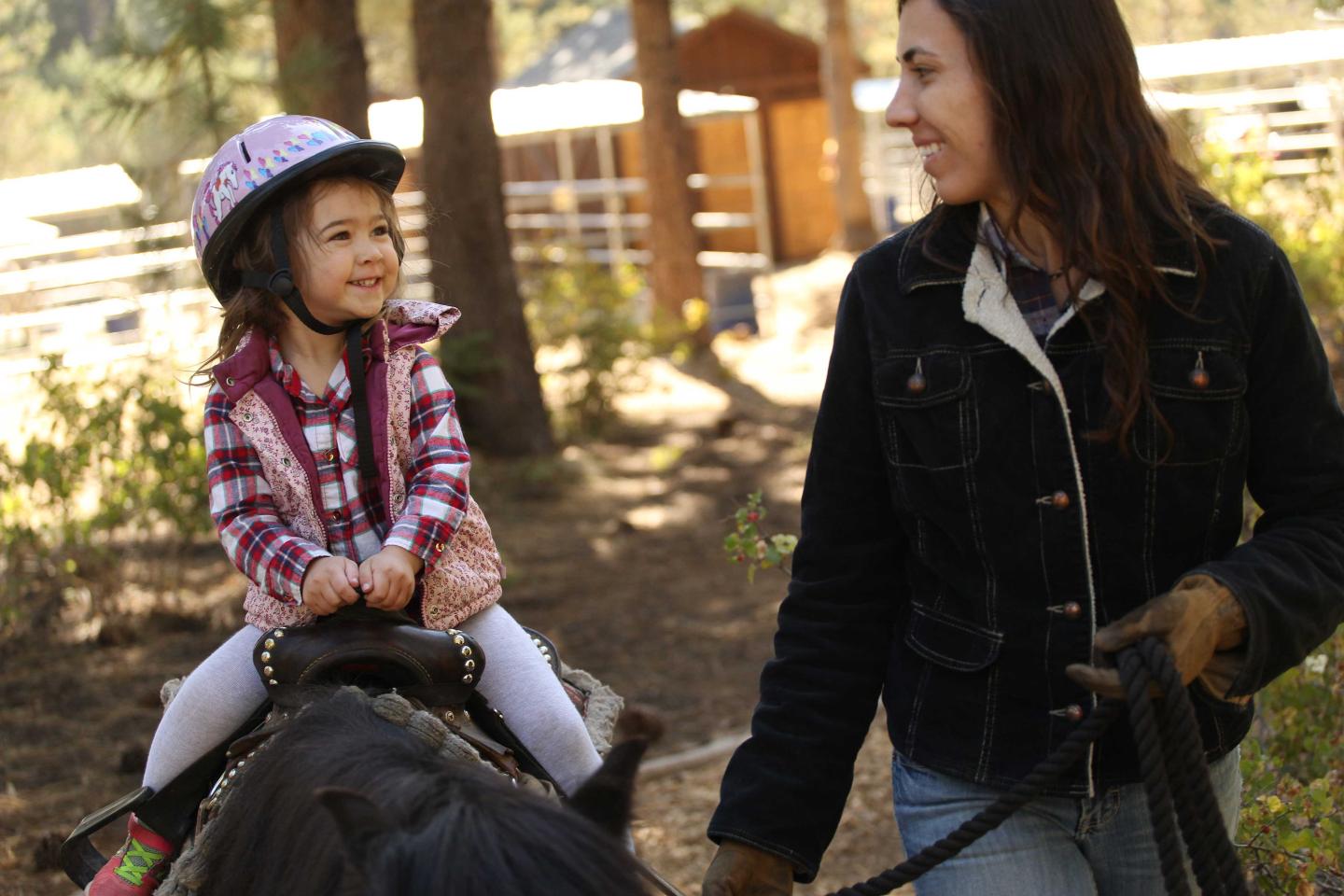 Child on horse with helmet, smiling at woman in outdoor setting.