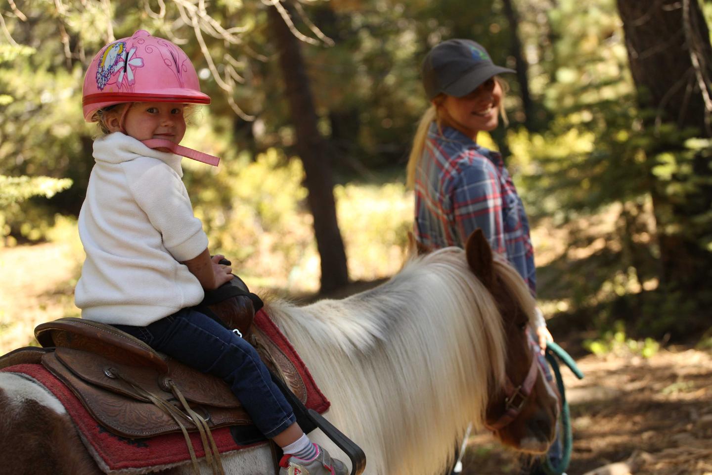 Child in pink helmet riding a pony, guided by a woman in a forest setting.