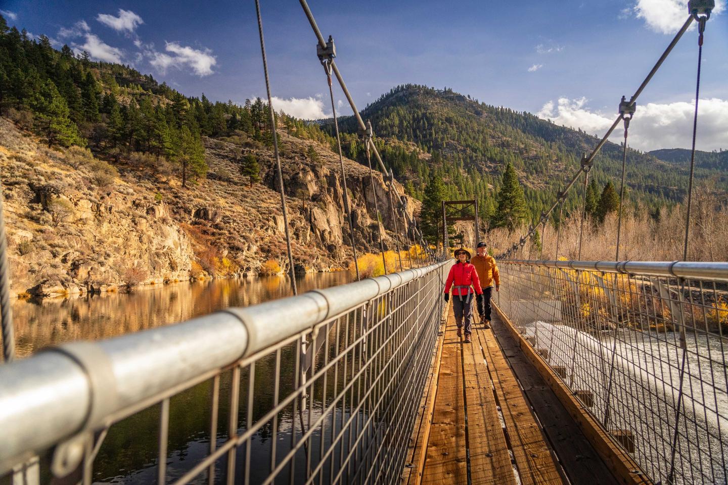 Two people walking on a wooden suspension bridge in a mountainous landscape.