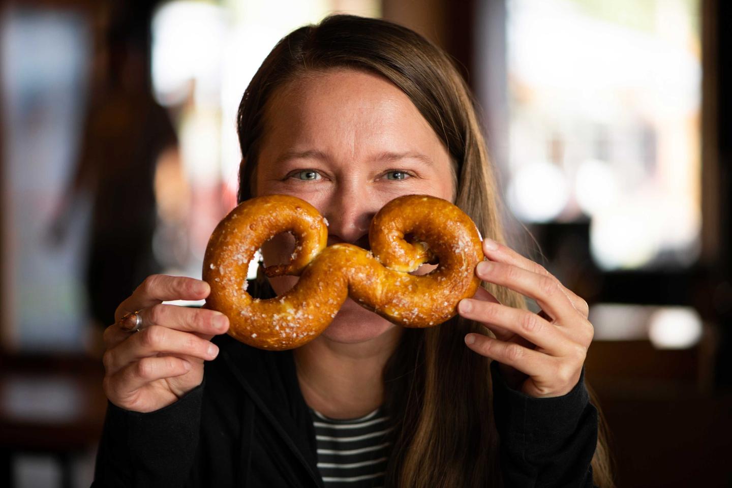Woman playfully holds pretzel over her face in a cozy setting.