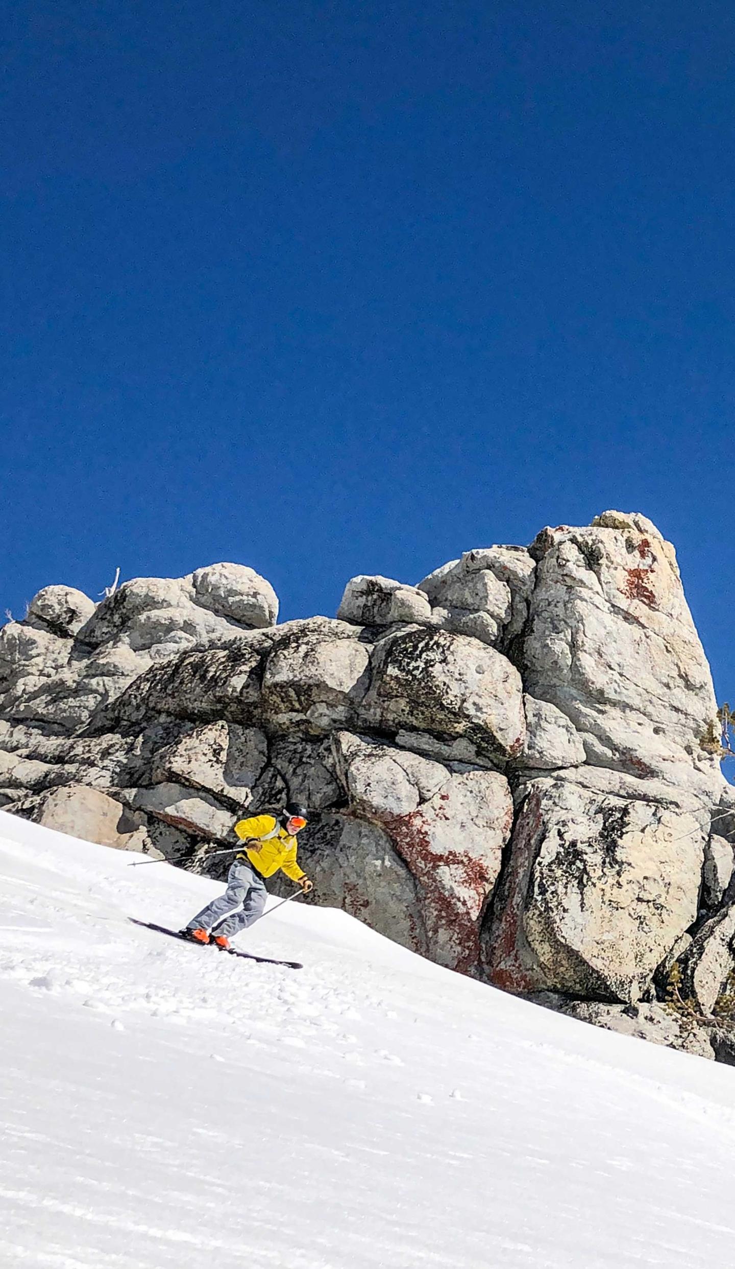 Skier in yellow descends snowy slope beneath blue sky and rocky outcrop.
