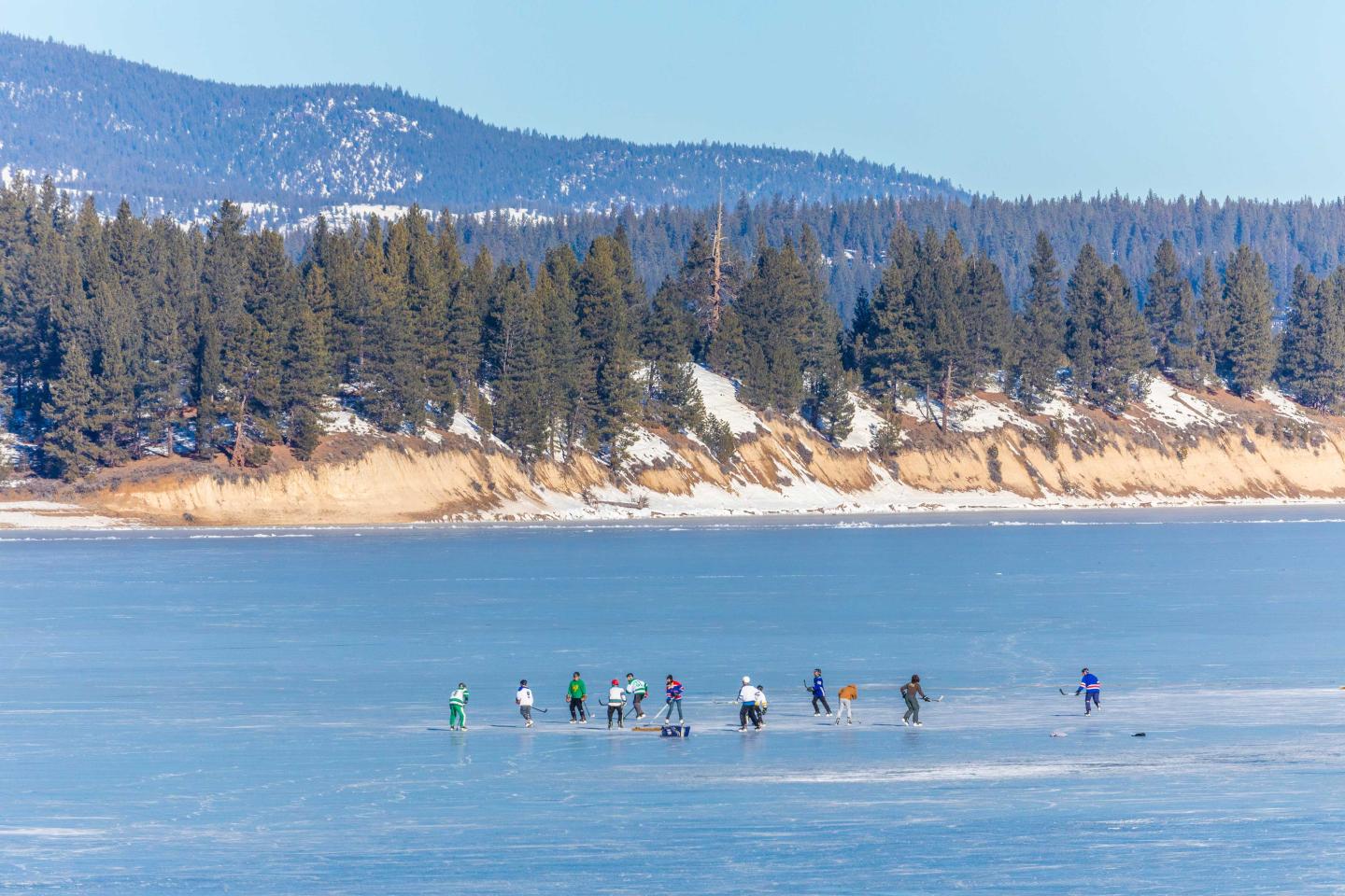 People skating on a frozen lake with snow-covered trees and mountains in the background.
