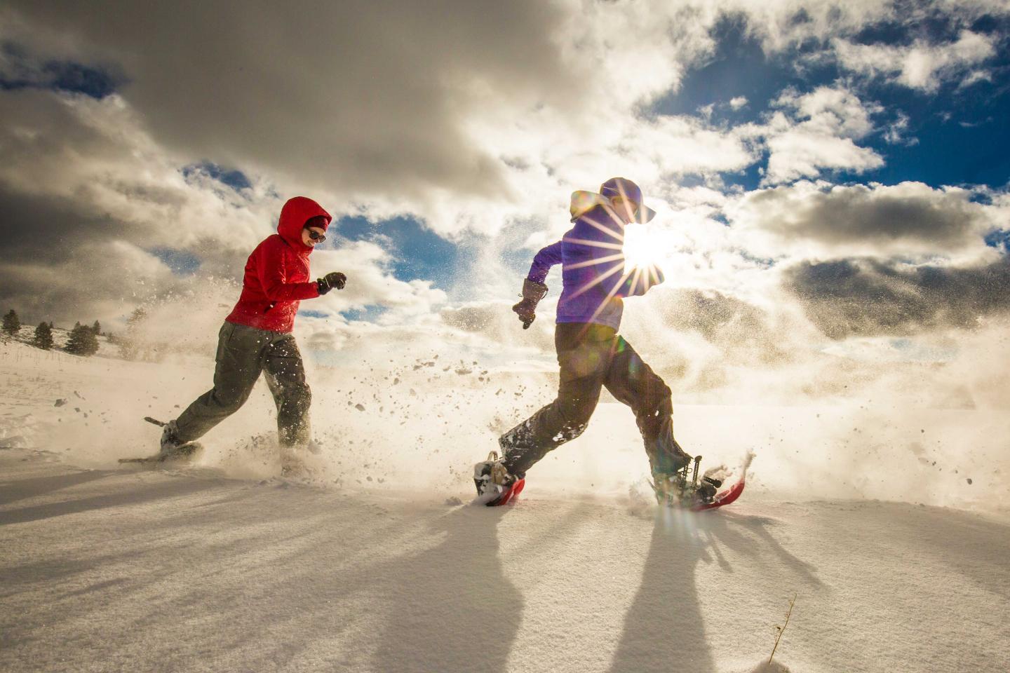 Two people snowshoeing under a cloudy sky with sun rays peeking through.