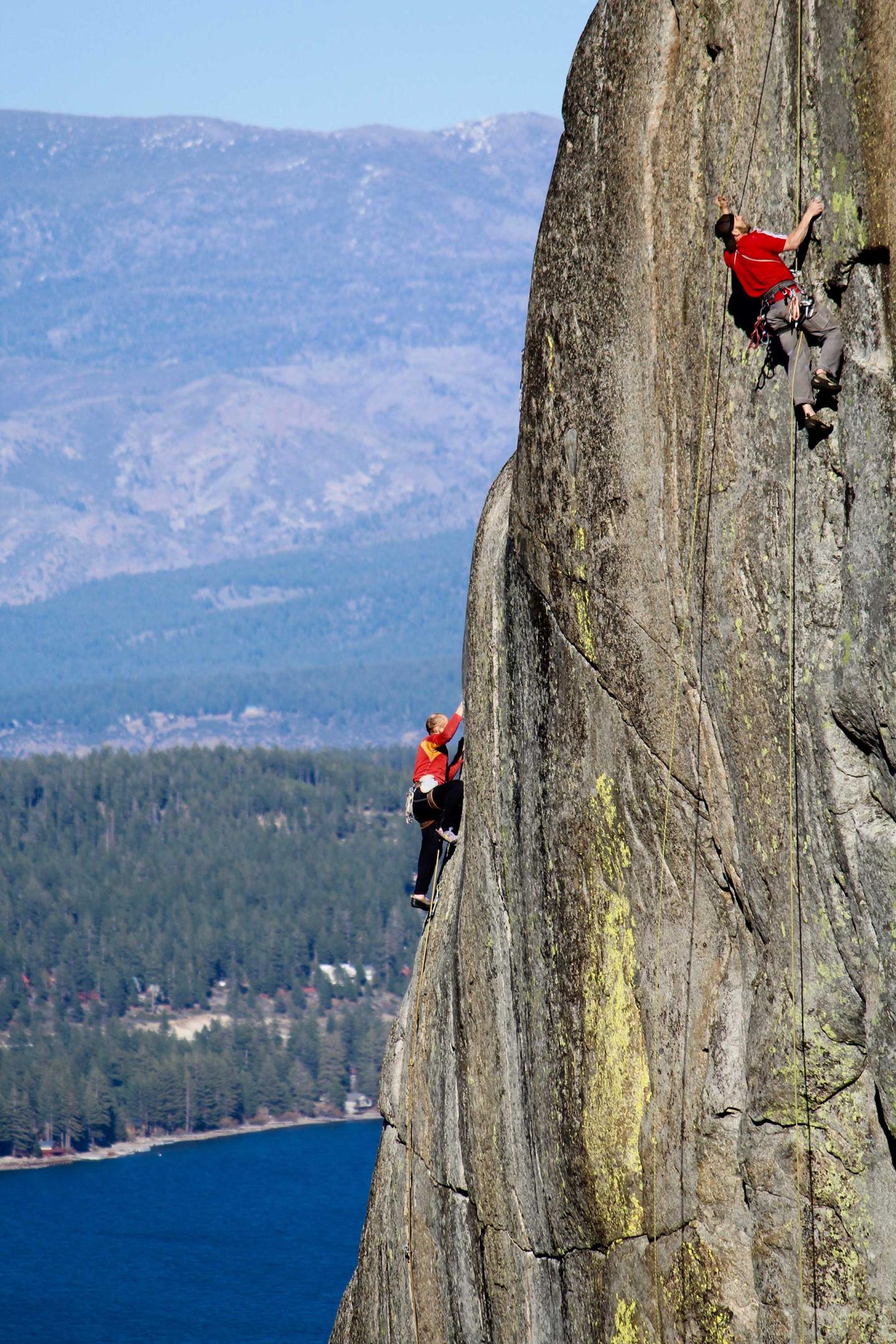 Climbers scaling a steep rock face with mountains and lake in the background.