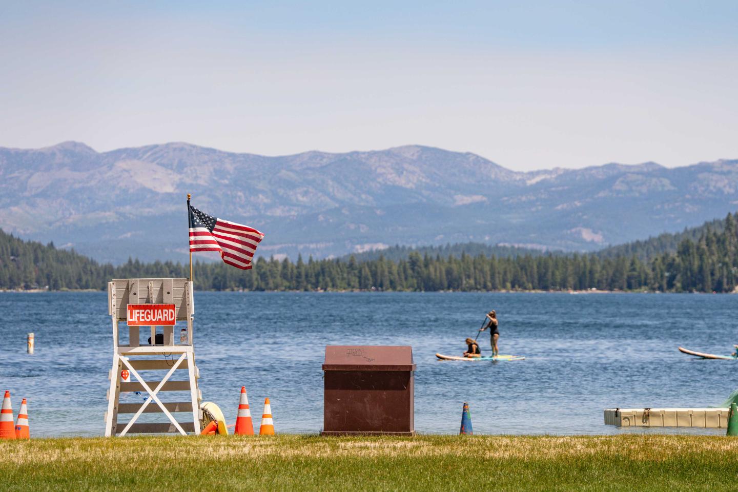 Lifeguard chair by lake with paddleboarders, mountains in background.