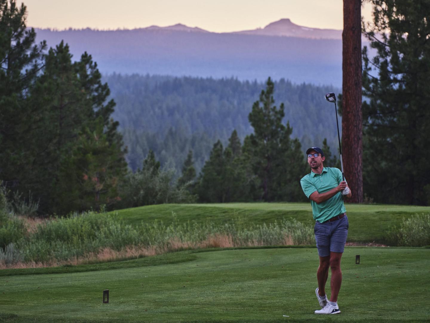 Golfer in mid-swing on a lush course, with mountains in the background at dusk.