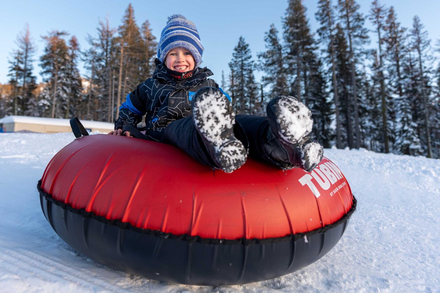 Child smiling on a red snow tube, surrounded by snowy trees.
