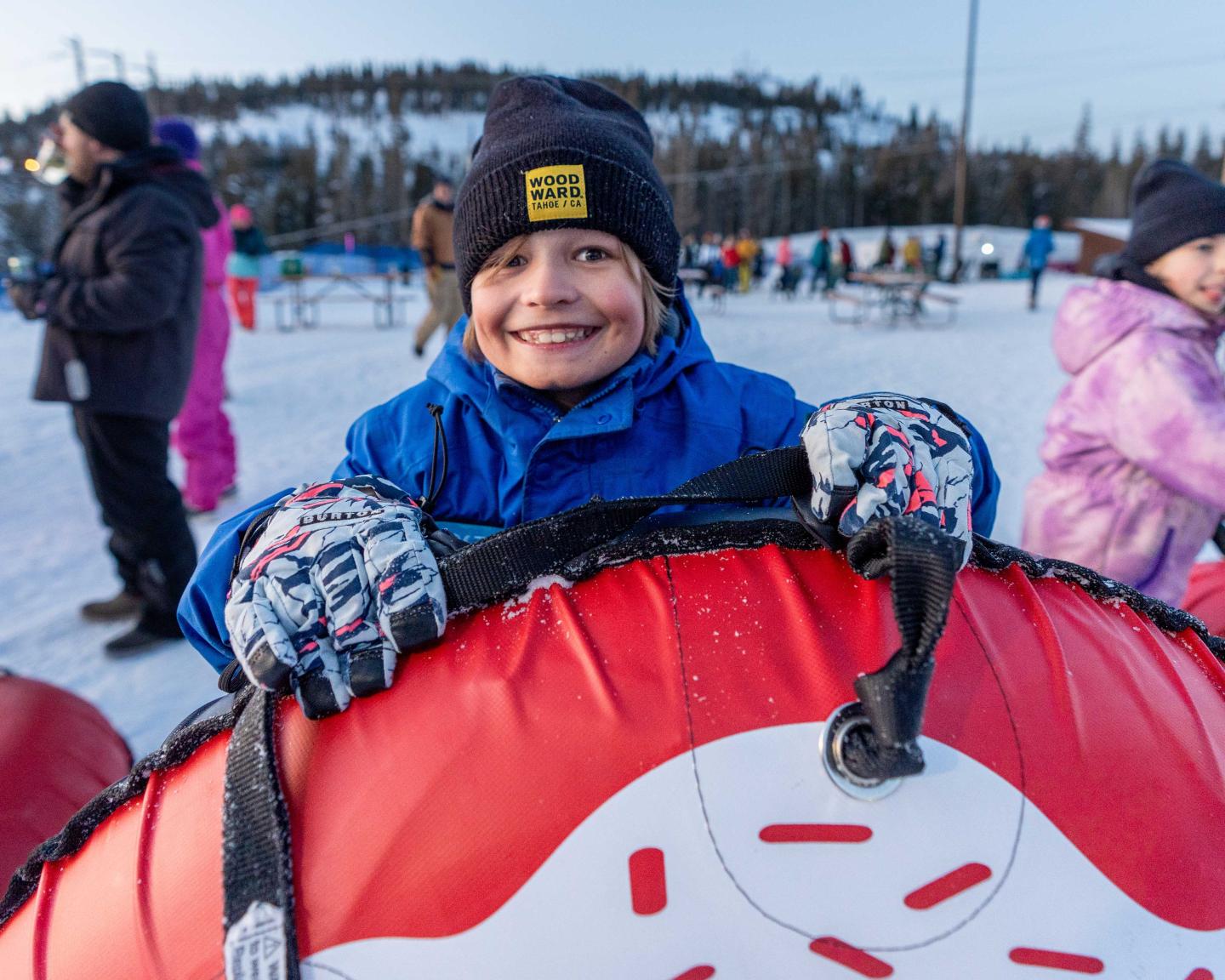 Smiling child in winter gear holds a snow tube, surrounded by snowy landscape.