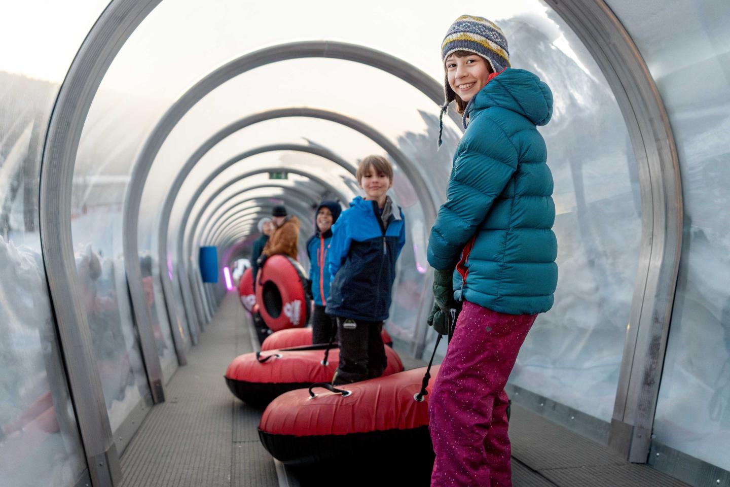 Children in winter clothing with snow tubes in an indoor ski tunnel.