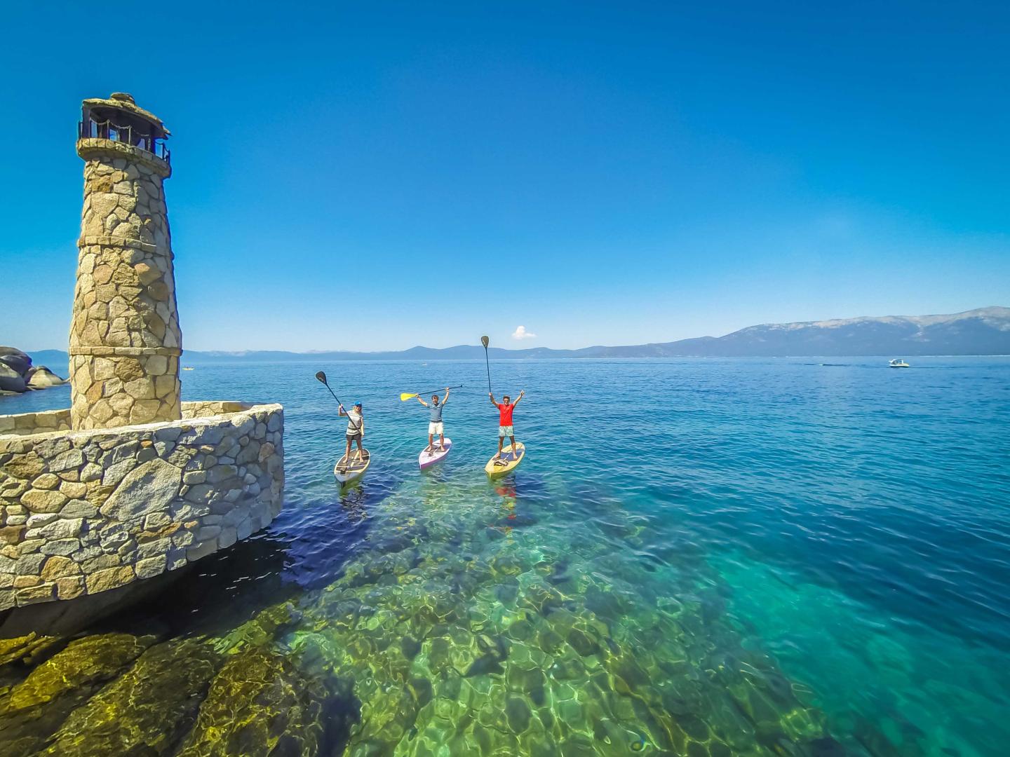 Three paddleboarders near a stone lighthouse on clear blue water.