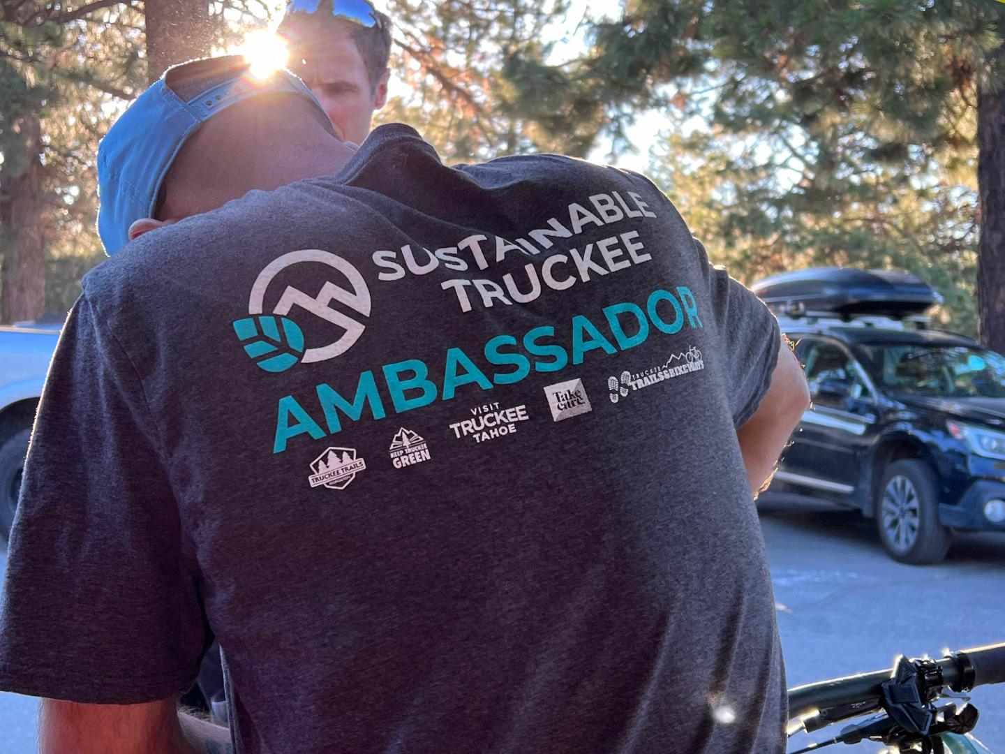 Man in a "Sustainable Truckee Ambassador" shirt outdoors near parked cars.