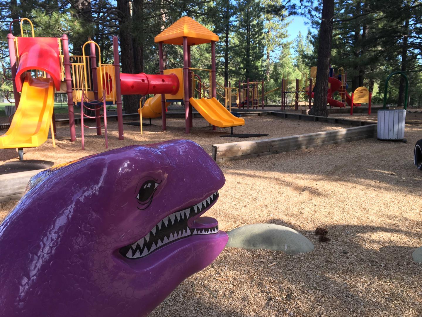 Purple dinosaur sculpture in a playground with slides and trees.