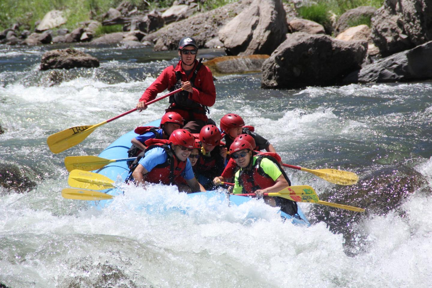 Group whitewater rafting, navigating rapids with paddles, wearing helmets and life jackets.