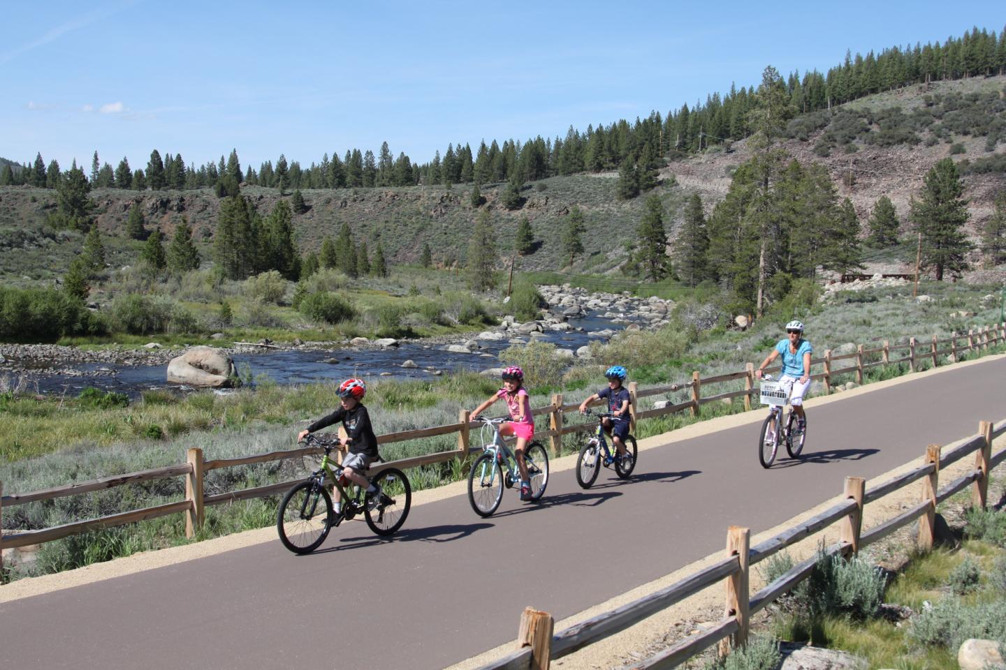 Four people cycling on a scenic path by a river and trees.