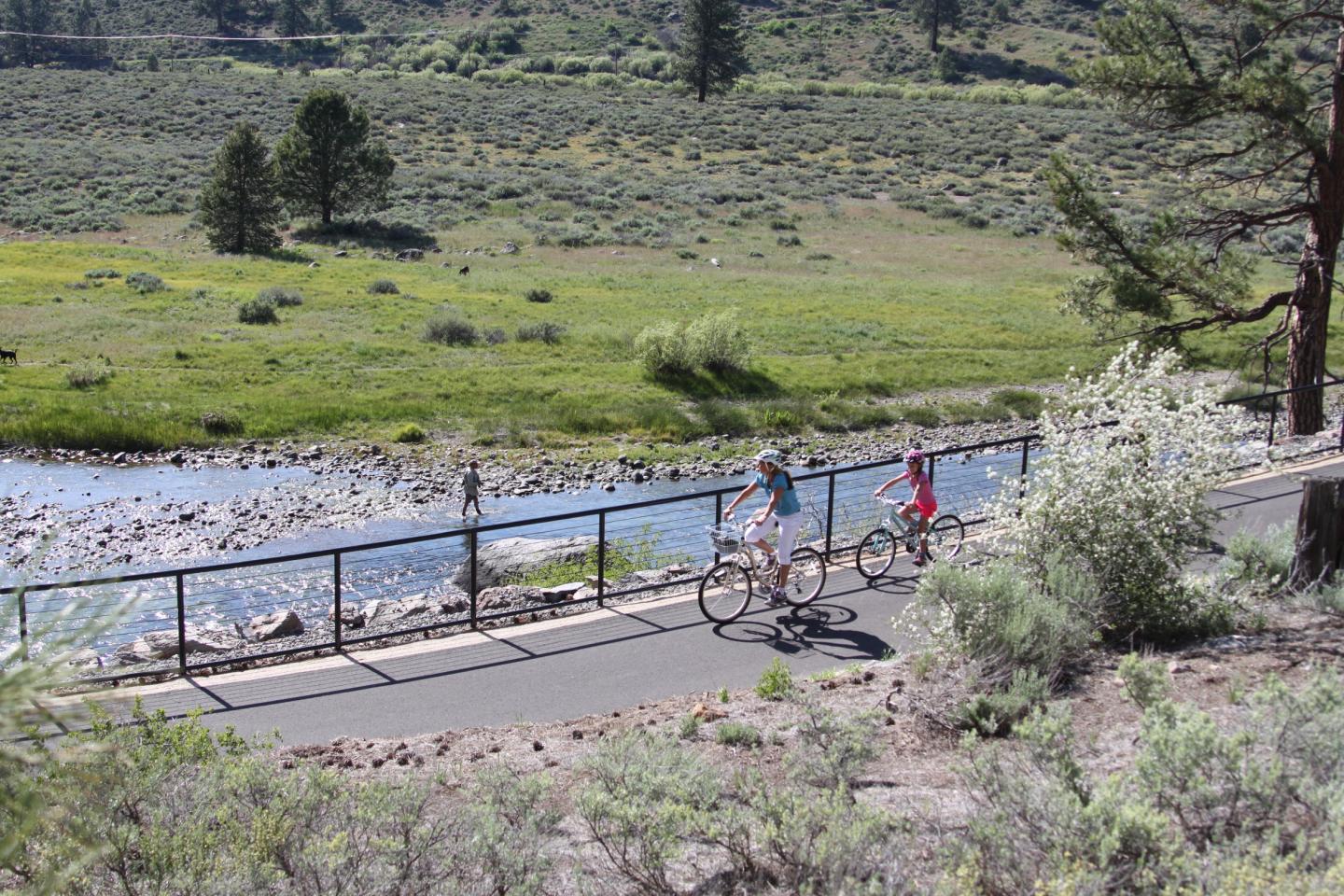 Cyclists riding along a riverside path in a scenic, grassy landscape.