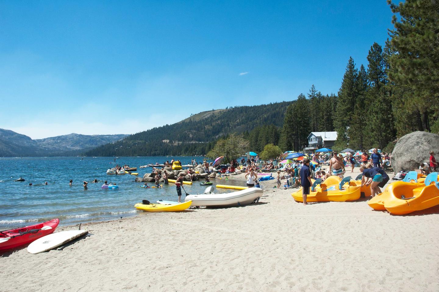 Crowded beach with kayaks and swimmers, surrounded by trees and mountains.