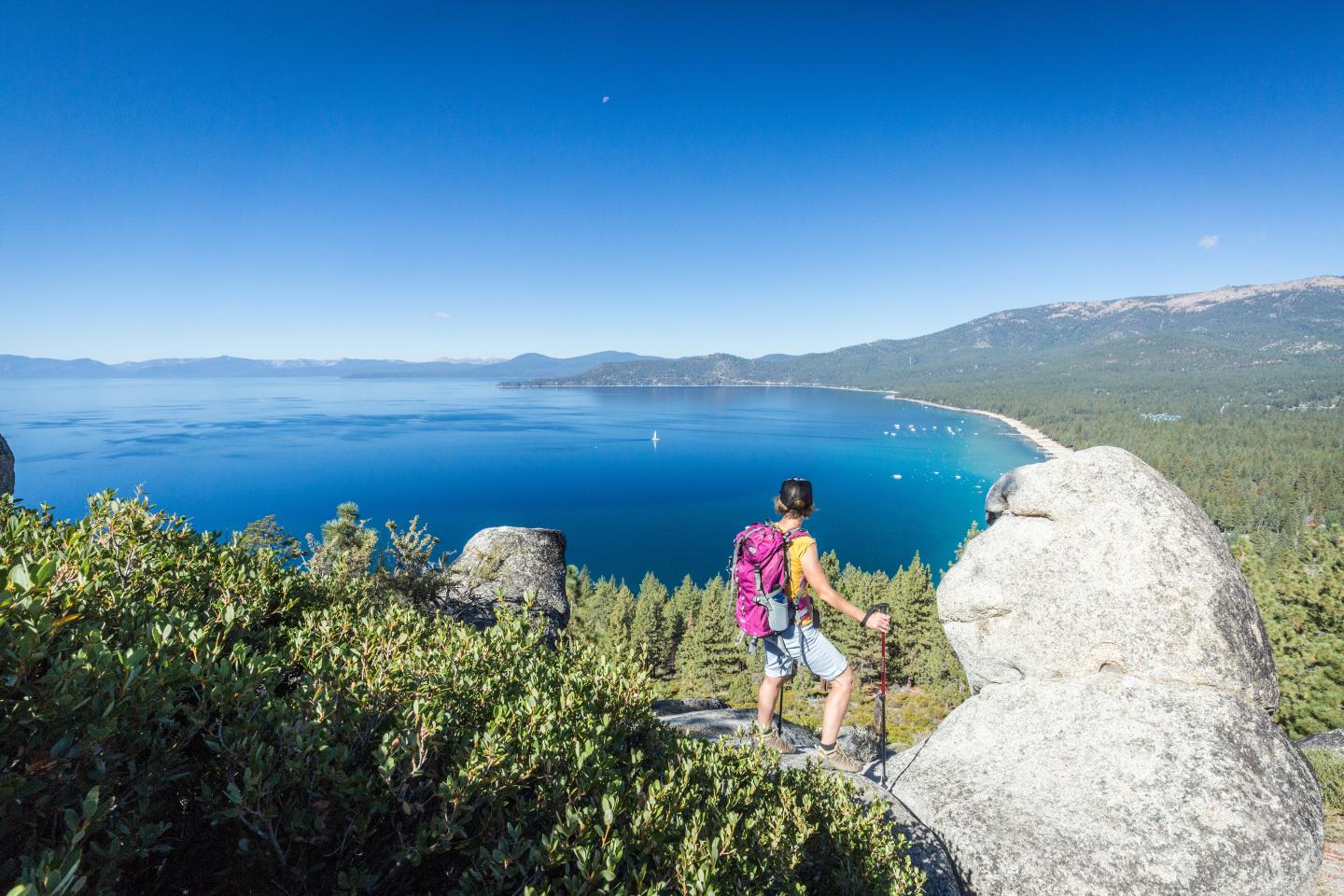 Hiker with a backpack overlooks a vast blue lake from a rocky hilltop.