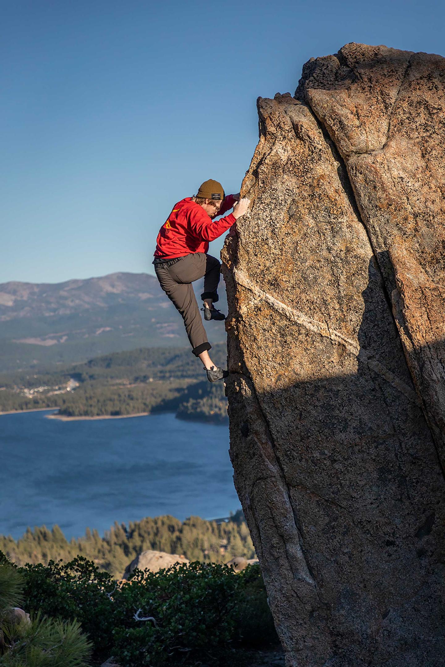 Climber scaling large rock, lake and mountains in background.