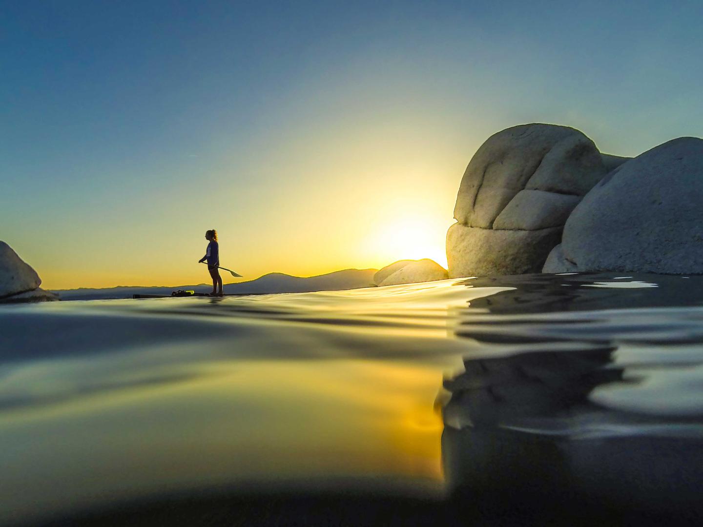 Sunset over water with person walking, waves and large rocks nearby.