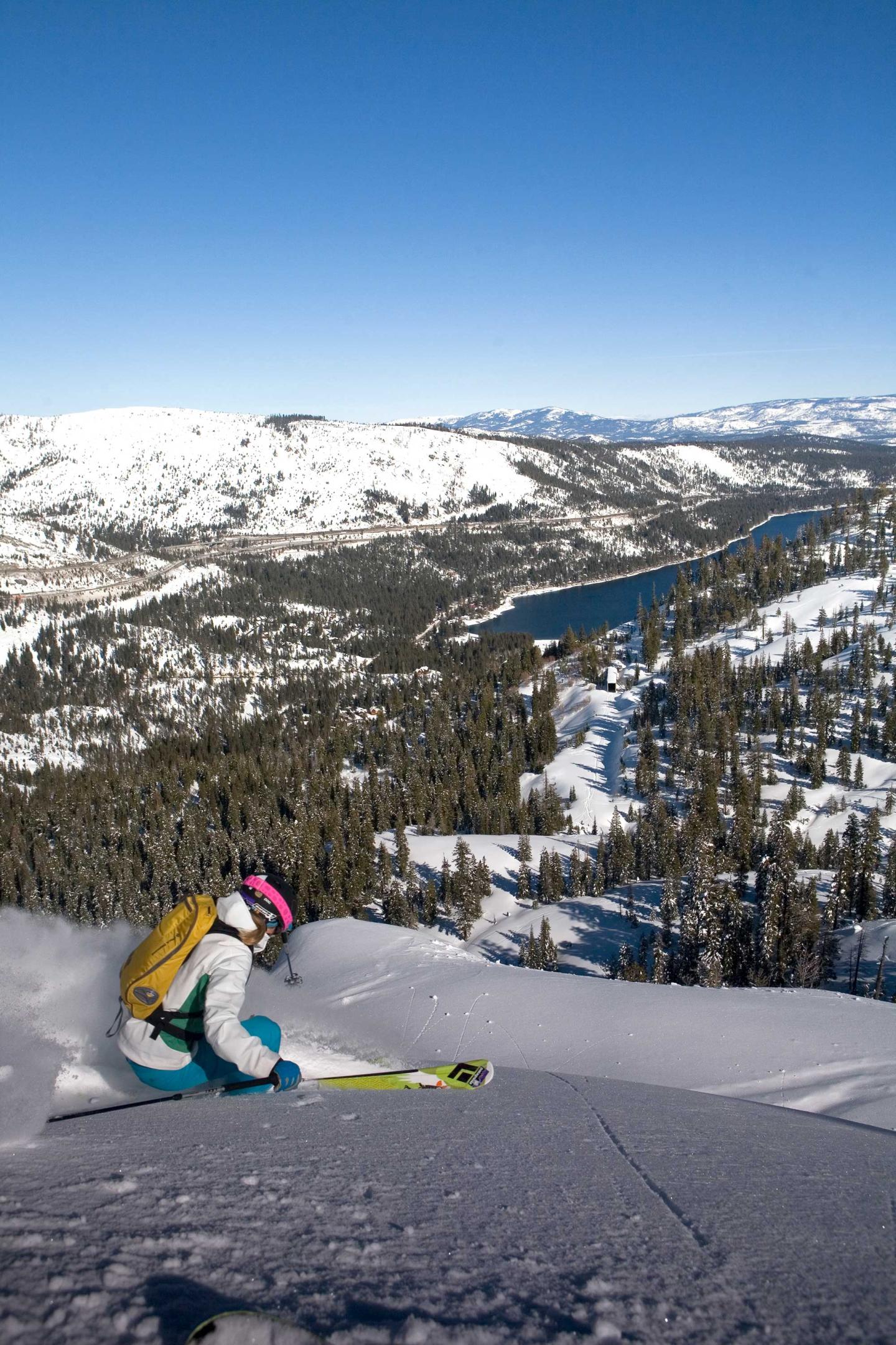 Skier in colorful gear on snowy mountain, overlooking a forest and lake in distance.