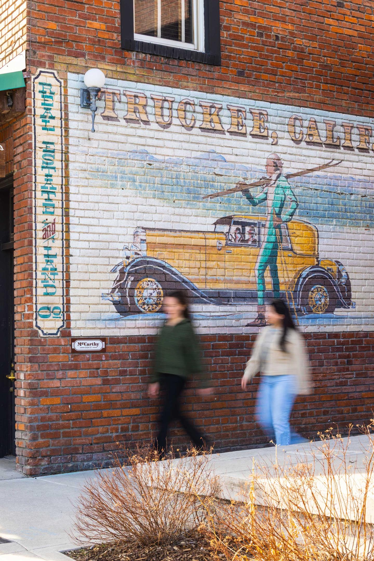 Brick wall mural with a vintage car and two people walking in foreground.