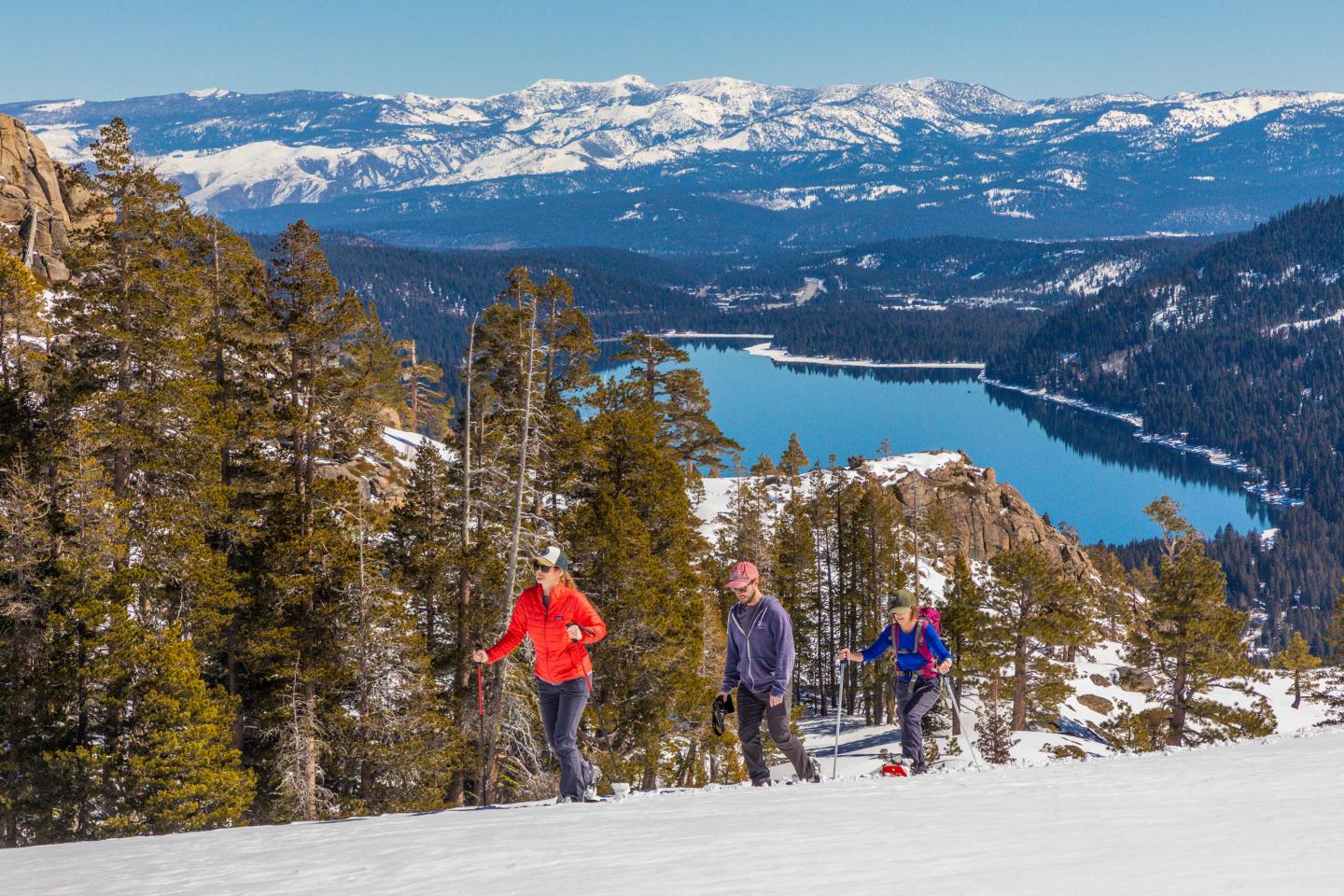 Three people hiking on a snowy mountain trail with a lake and mountains in the background.