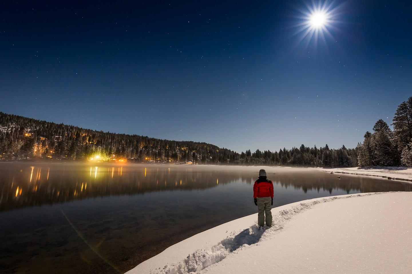 Person in red jacket stands on snowy shore under moonlit sky by a calm lake.
