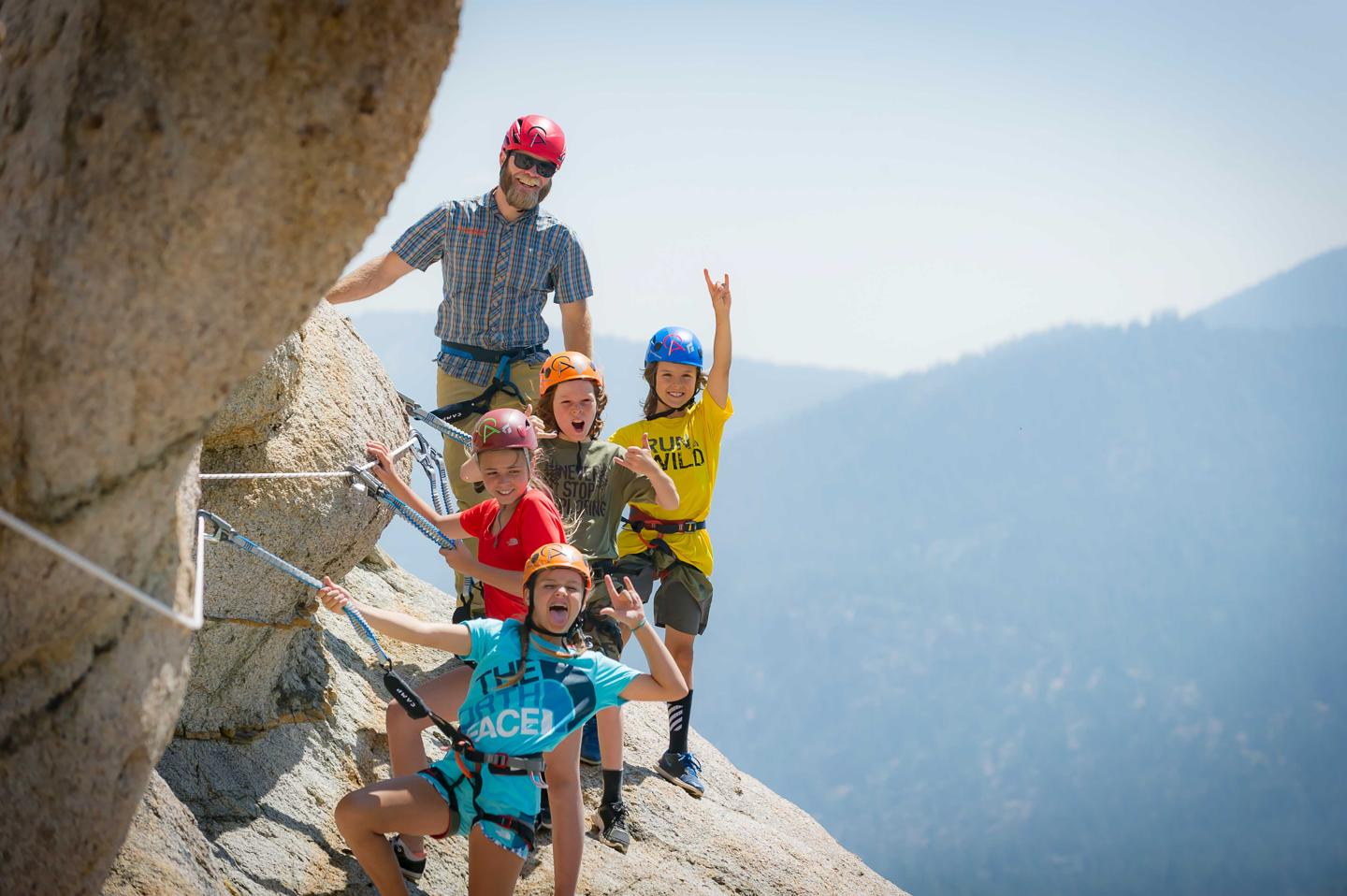 Five climbers smiling on a mountain ledge, wearing helmets and harnesses.