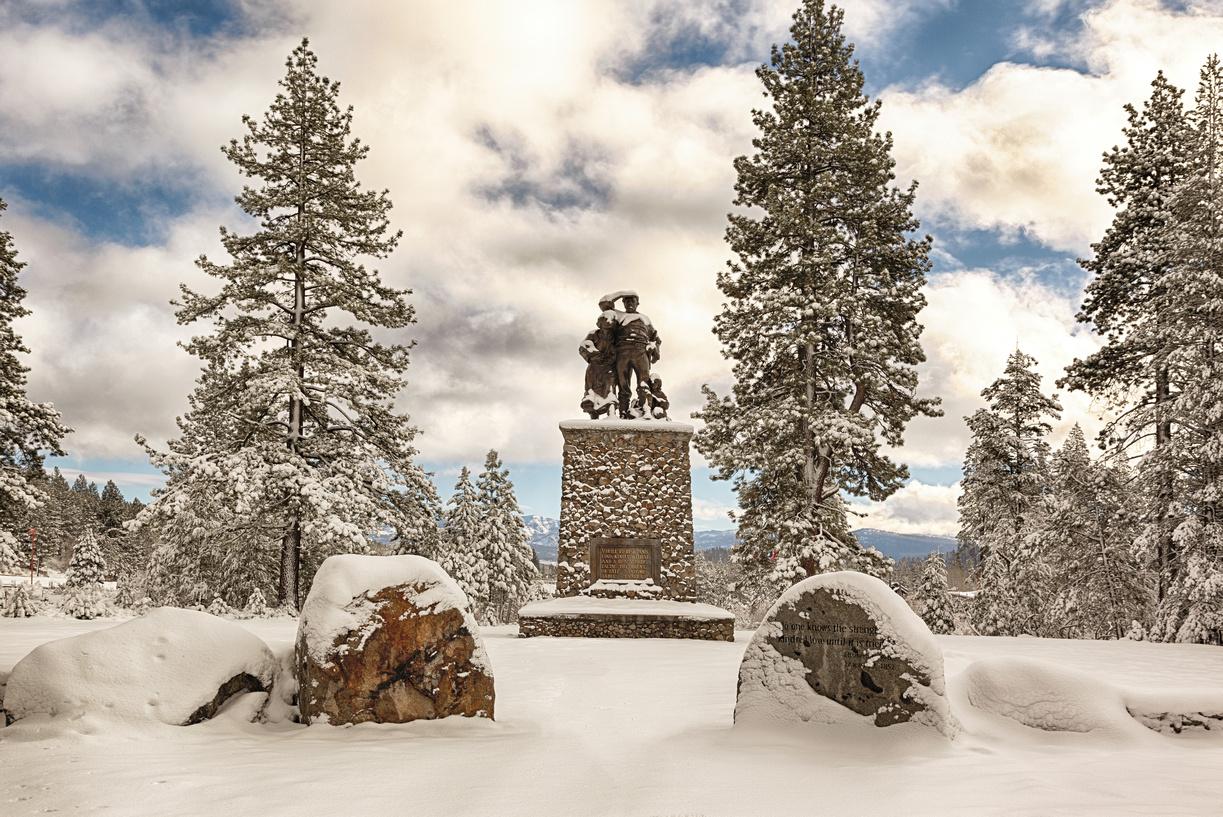 Snow-covered statue surrounded by tall trees under a cloudy sky.