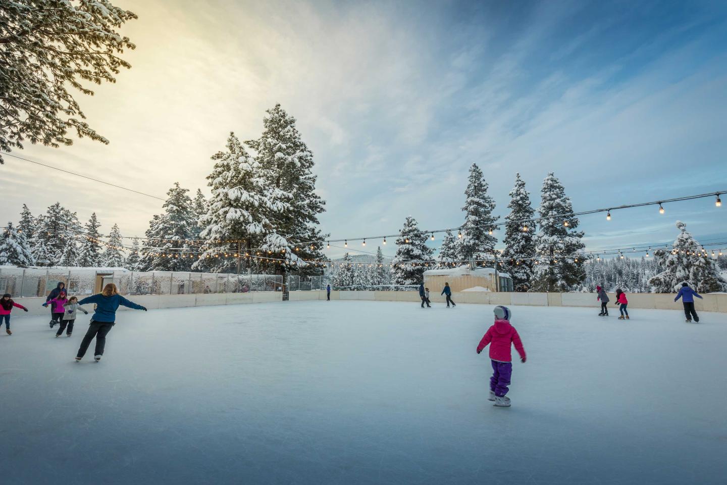 Snowy outdoor rink with people ice skating; sun setting behind tall trees.
