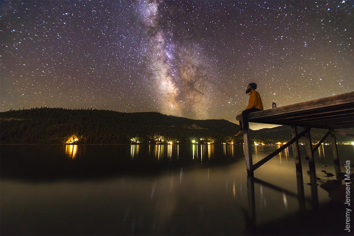 Starry sky over a lake, person sitting on a dock gazing at the Milky Way.
