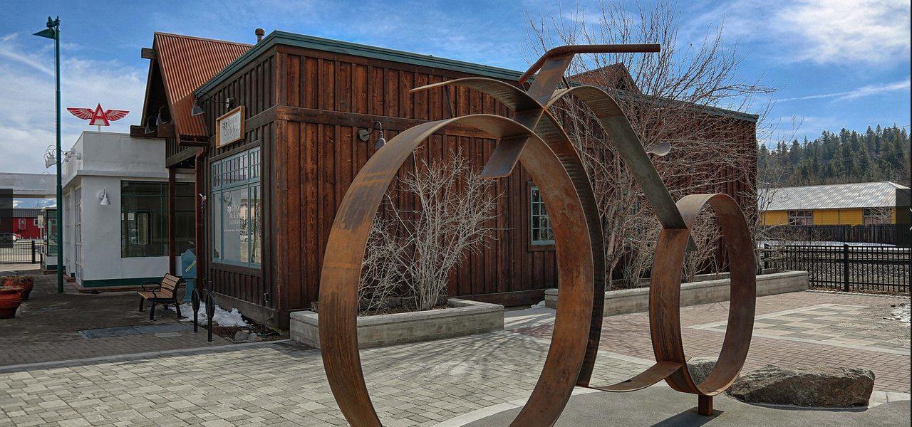 Large abstract metal sculpture in front of a wooden building on a sunny day.