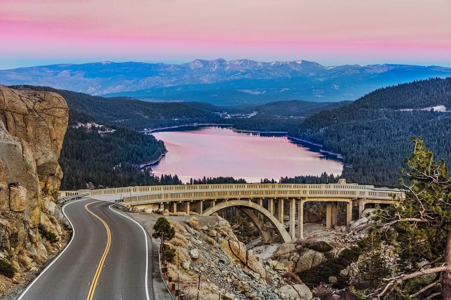 Curving road and bridge over a scenic lake at sunset with mountains in the distance.