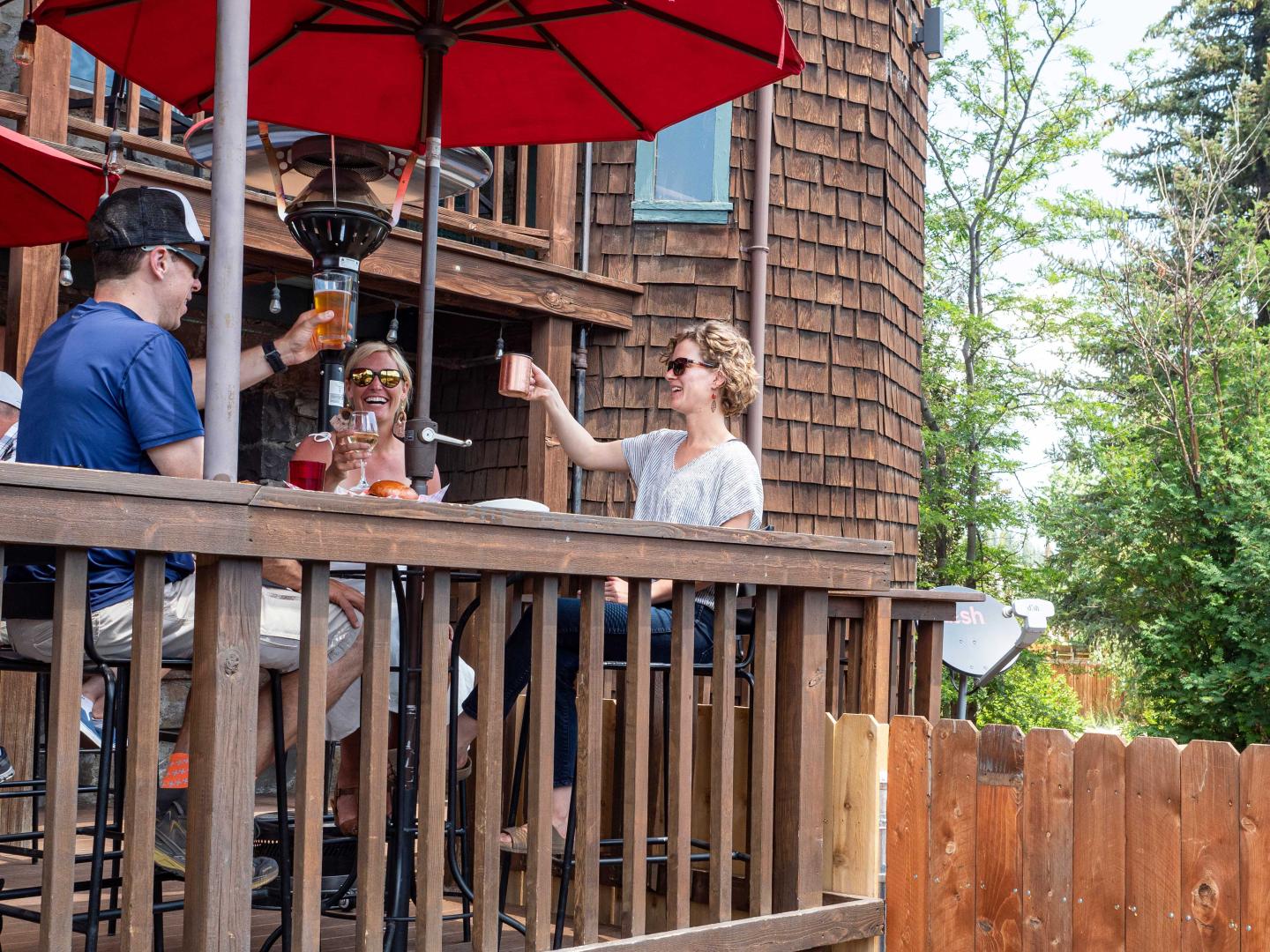 People toasting drinks under red umbrellas on a wooden deck.