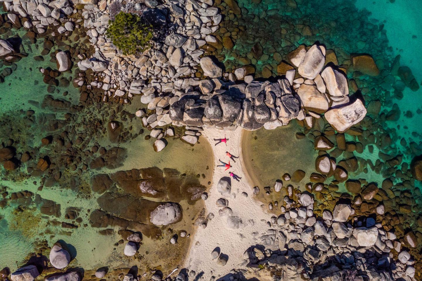 Aerial view of a rocky beach with two people lounging on sand, surrounded by turquoise water.