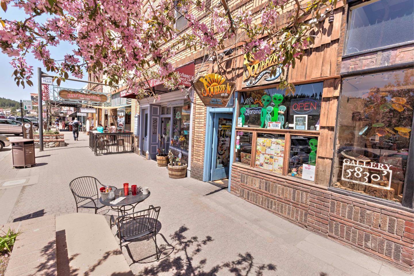 Street with colorful storefronts and blooming pink tree, outdoor seating visible.