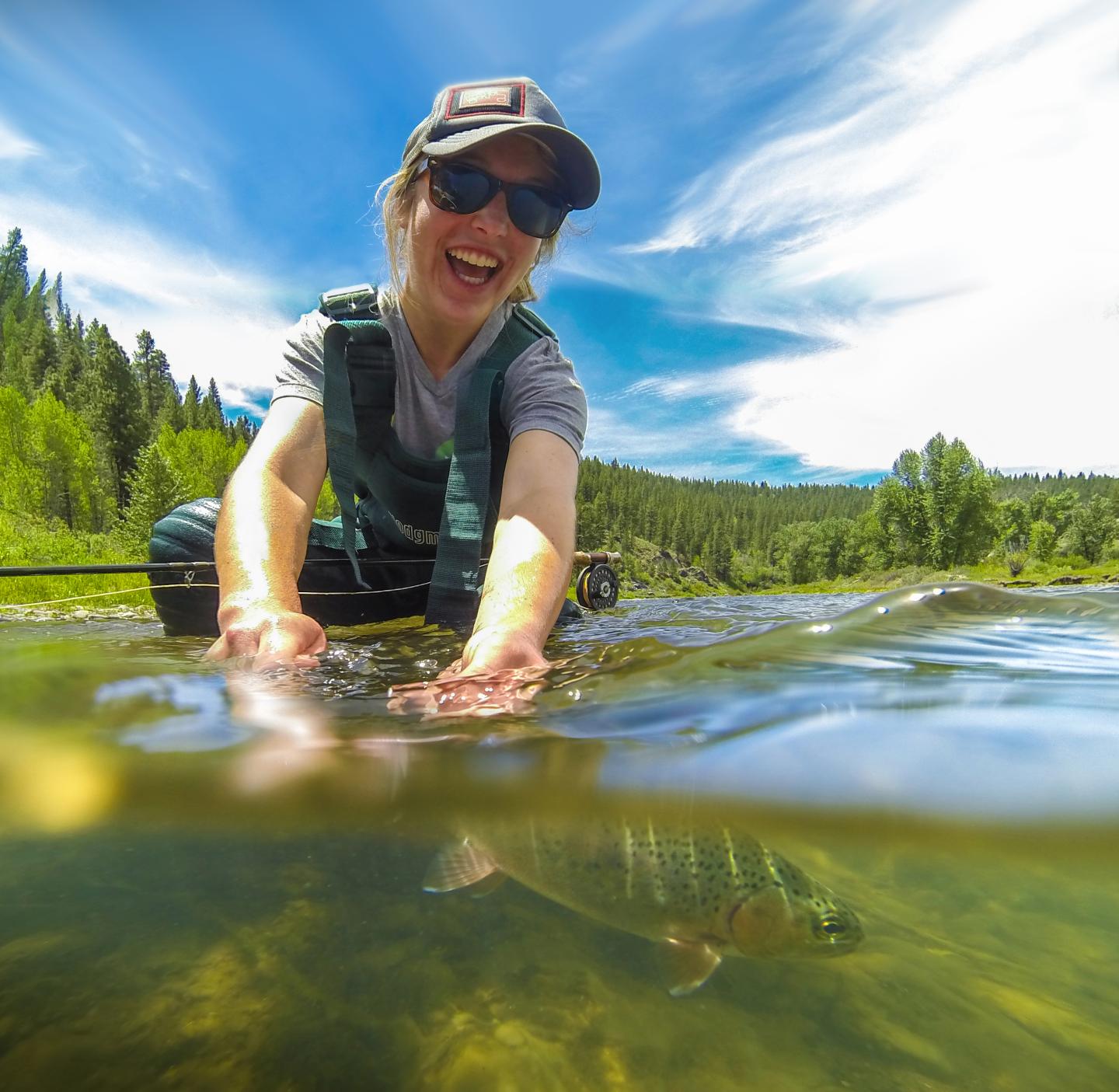 Person smiling while releasing a fish into a clear river under a sunny sky.