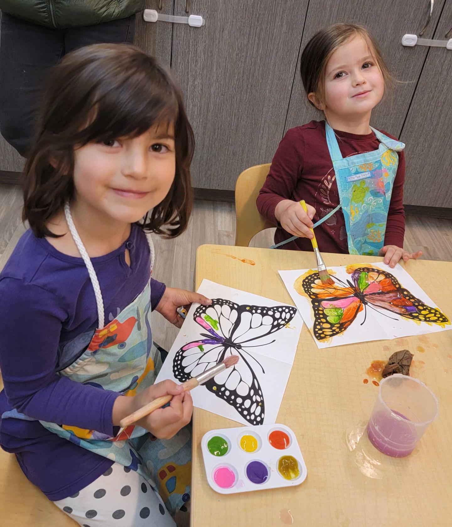 Young children painting butterfly illustrations at a table, smiling.