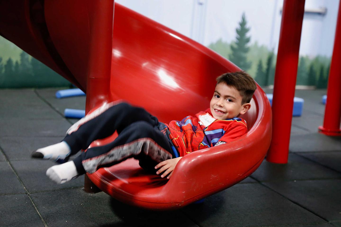Child sliding down a red playground slide, smiling, wearing a red sweatshirt.