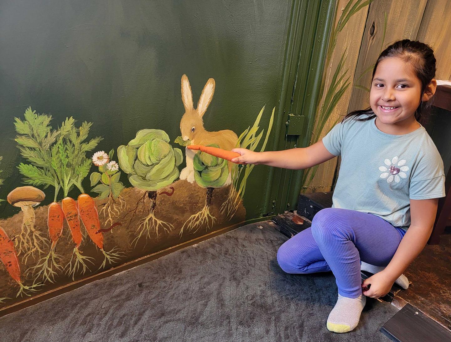 Child smiling and holding a carrot to a bunny mural on a wall.