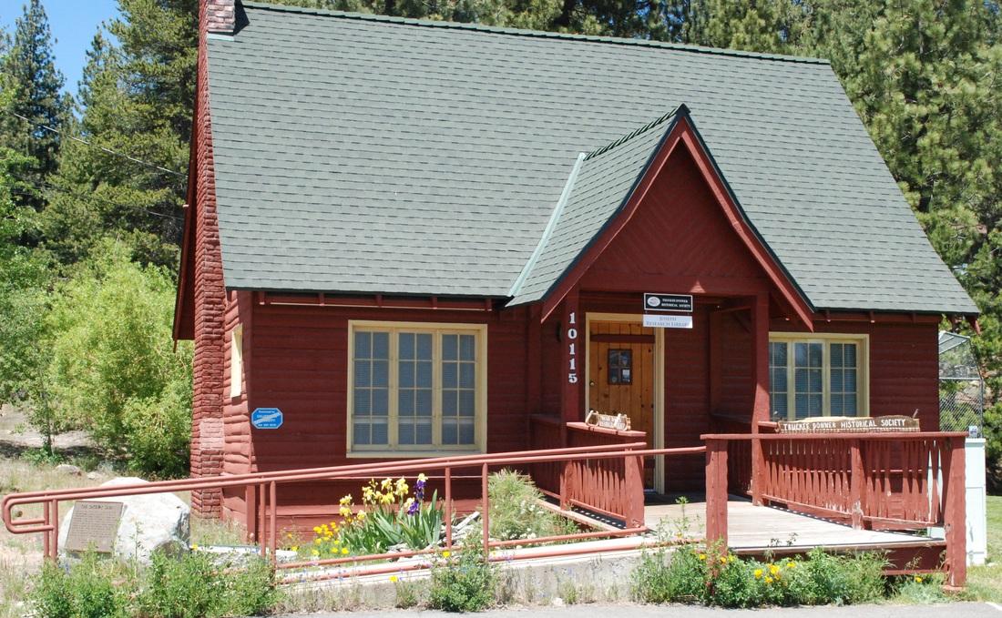 Red cabin with green roof surrounded by trees and flowers.