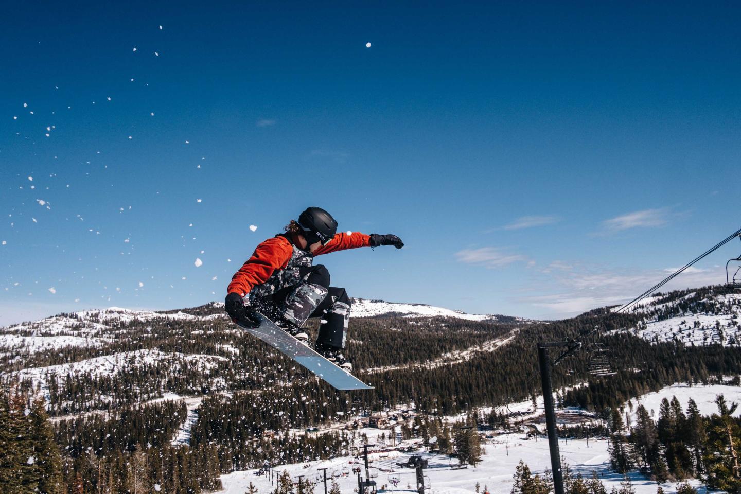 Snowboarder in mid-air jump, snow-covered mountains, clear blue sky.