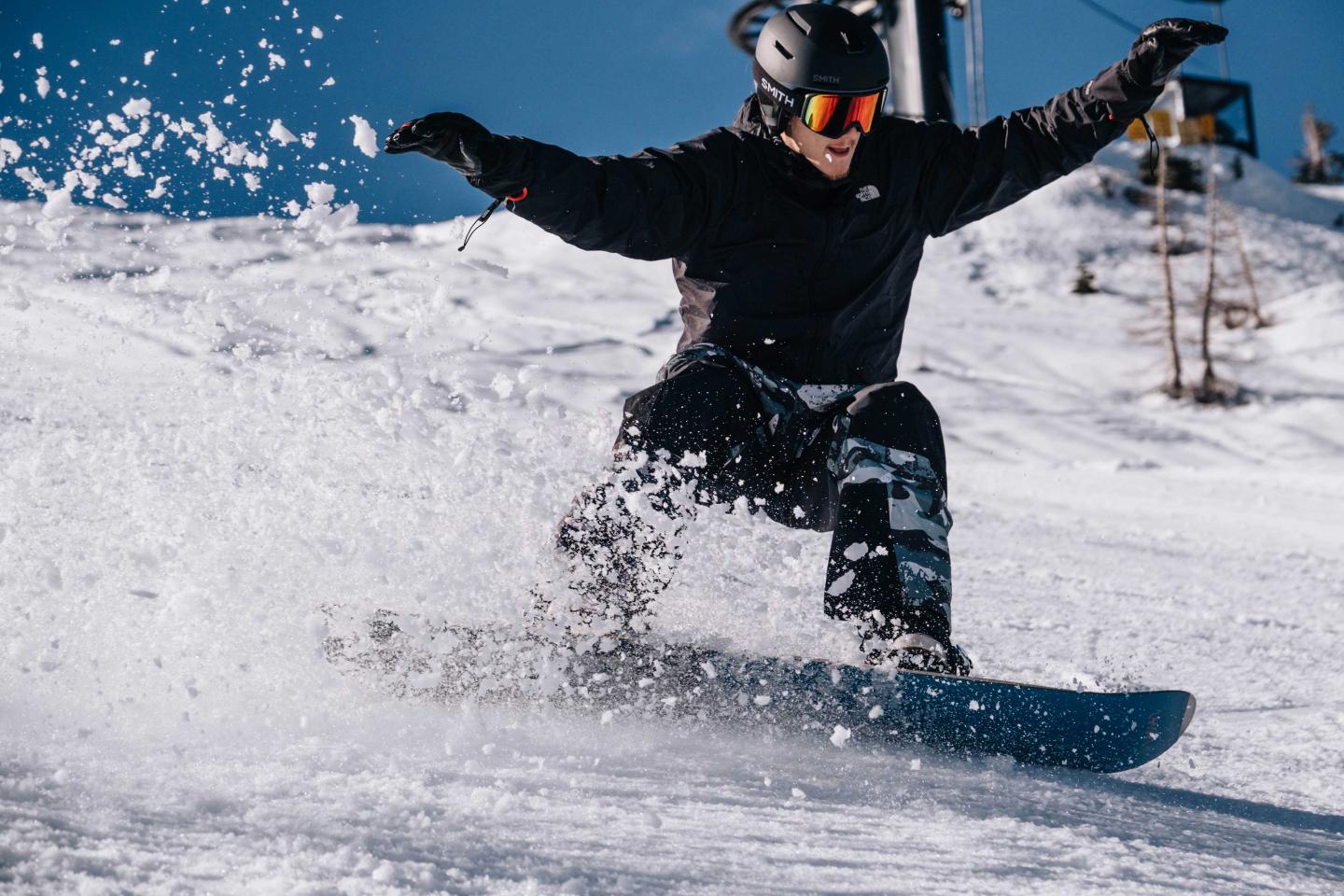 Snowboarder carving through snow on a sunny day, wearing black gear and goggles.