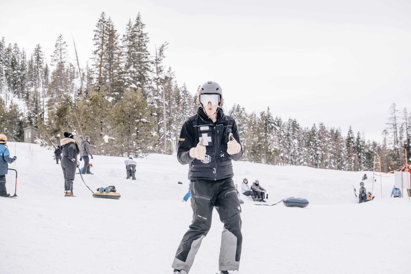 Person in ski gear giving thumbs up on snowy slope with trees in background.